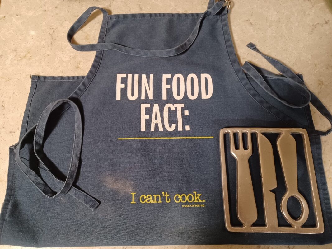 Photo of silver metal trivet in the shape of a flattened fork, knife, and spoon on top of a blue apron that reads Fun Food Fact: I can't cook.
