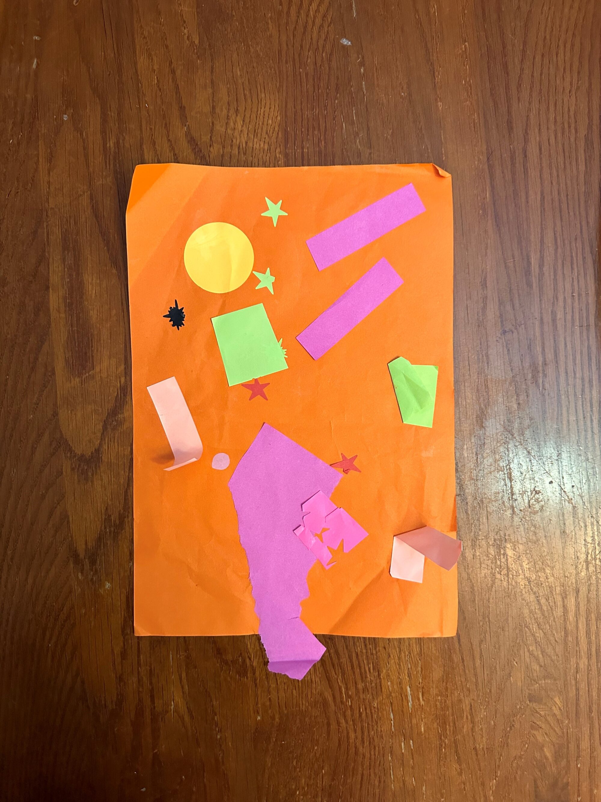 Photo of an orange piece of construction paper with cut paper shapes in various colors pasted on top.
