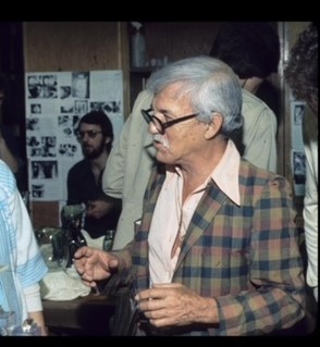 Photo of an elderly man wearing dark, thick-rimmed glasses, pink button-down shirt, and plaid jacket.