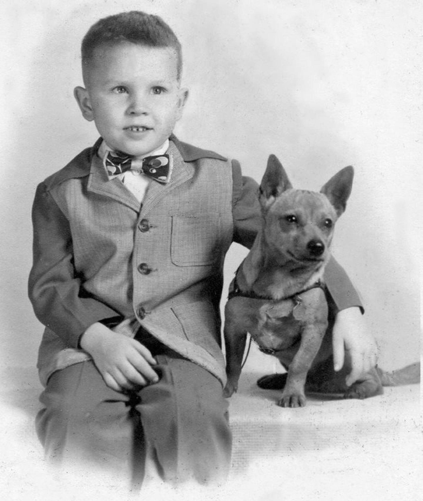 Black and white photo of a young boy in a suit with his arm around a small dog.