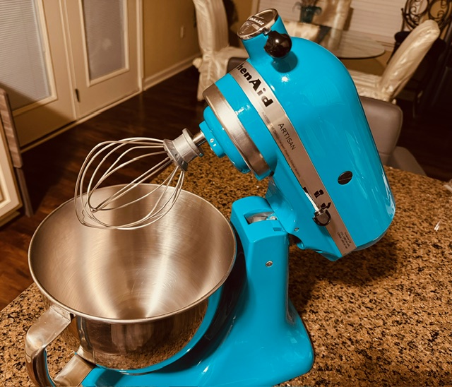 Photo of a blue stand mixer with a stainless steel mixing bowl on top of a stone countertop.