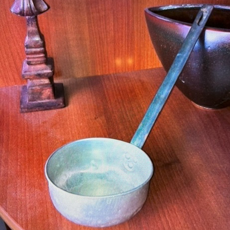 Photo of an antique metal ladle sitting on a wooden table next to a bowl and candlestick.